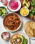 Taco ingredients including pulled pork, pickled onions, lime wedges, pineapple, avocado, radishes, and cilantro.