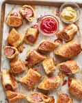 Pigs in a blanket on a baking sheet with bowls of ketchup and mustard.