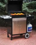 Outdoor gas grill with open lid, grilling orange and yellow peppers, set on a brick patio with evergreen trees in the background.
