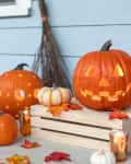 Carved pumpkins with candles and autumn leaves on a porch, next to a broom and wooden crate.