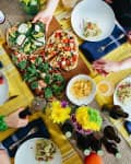 People sharing a meal with salad, pizza, chips, and drinks on a yellow-striped tablecloth, surrounded by flowers.