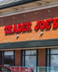 Trader Joe's storefront with red signage on a brick building, a woman with a shopping cart, and a parked red car.