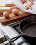 A cast iron skillet is seen on top of an electric stove, while eggs, butter and meat is placed on a wooden chopping board besides it