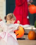 Toddler in a pink dress holding a pumpkin bucket, walking with an adult during Halloween.