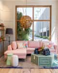 Living room with pink sectional sofa, geometric coffee table, colorful pillows, and large window with plants outside.