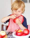Child in a red bib holding an apple, seated at a table with more apples in a bowl.