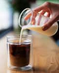 Pouring milk into a glass of iced coffee on a wooden table.