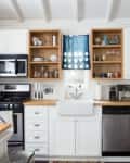White kitchen with stainless steel appliances, open shelving, farmhouse sink, and blue curtain above the window.