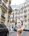 Person in shorts and hat walking down a narrow Parisian street lined with ornate buildings and parked cars.