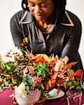 Woman arranging a vibrant autumn floral centerpiece on a set dining table with wine glasses.