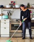 Woman mopping kitchen floor with white cabinets, stainless steel appliances, and open shelves with dishes.