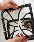 Hands washing a black stove grate under running water in a white sink.