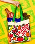 Hand holding a Trader Joe's tote bag filled with groceries, including a baguette and leafy greens, against a dollar bill pattern.