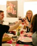 Friends toasting with wine glasses at a dinner table with a pink striped tablecloth, flowers, and candles.