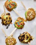 Caramel apple slices on sticks topped with candy, nuts, and cookie crumbs on a parchment-lined surface.