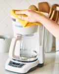 Hand cleaning a white coffee maker with a yellow cloth on a kitchen counter.