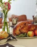 Roasted turkey with apples and sage on a table set with flowers, glassware, and bread rolls.