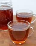 Two glass mugs and a jar filled with amber-colored tea on a wooden surface.
