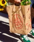 Person carrying Trader Joe's paper bag with sunflowers and greens, wearing jeans and green sneakers.