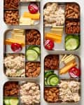 Four meal prep containers with chicken salad, almonds, pecans, cheese slices, cucumber, and Babybel cheese.