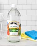 A bottle of Heinz distilled white vinegar on a kitchen counter next to sponges, a cleaning cloth, and a spray bottle