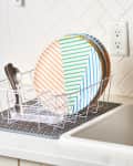 Dish rack with striped plates and cutlery beside a sink, soap dispenser, and herringbone tile backsplash.