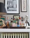 Home bar setup on a white shelf with various liquor bottles, a black lamp, framed art, and a vase of white flowers.