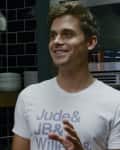Man in a kitchen with black tiled walls, wearing a white shirt with text, standing near a stack of plates and a microwave.