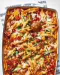 Baked penne pasta with meatballs, melted cheese, tomato sauce, and fresh parsley in a casserole dish.