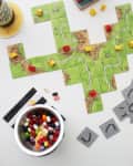Board game with green tiles and colored pieces, next to a bowl of jelly beans and a can on a coaster.
