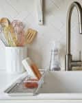 Kitchen sink with stainless steel faucet, white backsplash, dish soap, scrub brush, and utensils in a holder.