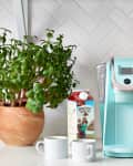 Mint green coffee maker on a kitchen counter with milk carton, two mugs, a plant, and a toaster.