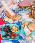 Child reaching for sliced bread on a picnic blanket with fruits, sandwiches, and a wicker basket.