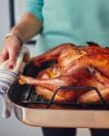 Person holding a roasted turkey in a metal roasting pan with striped towels.