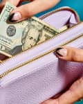 Hands placing a $20 bill into a lavender wallet with gold zippers against a blue background.