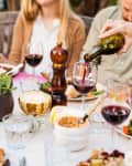 Outdoor dining table with people, wine glasses, candles, and plates of food, including vegetables and a pepper grinder.