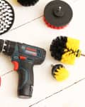 Cordless drill with various cleaning brush attachments in red, yellow, and black on a white wooden surface.