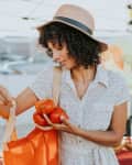 Woman in a hat placing tomatoes into an orange tote bag at a farmers market.