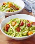 Zucchini noodles with cherry tomatoes, basil, and parmesan in white bowls on a wooden table.