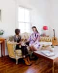 Two women sitting on a vintage sofa with a dog, next to a wooden coffee table and a guitar in a bright living room.