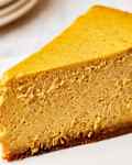 A slice of pumpkin cheesecake with the rest of the pumpkin cheesecake in the background.