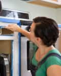 Woman painting kitchen cabinet white with a small roller, surrounded by appliances.