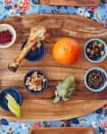 Wooden platter with an orange, bone, egg, artichoke, olives, nuts, and small bowls on a floral tablecloth.