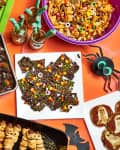 Halloween-themed snacks on a table, including mummy hot dogs, candy bark, apple teeth, and a magazine with pumpkins.