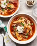 Two bowls of lasagna soup topped with ricotta, basil, and ground meat, next to a spoon and a bowl of pepper.