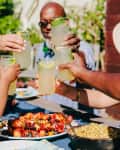 Group of people toasting with lemonade glasses over a table with grilled skewers and corn salad.