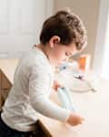 A young child in a white long-sleeve shirt is holding a blue towel while standing at a wooden kitchen counter with a glass bowl and ingredients nearby.