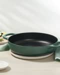 Green cast iron skillet on a table with sliced zucchini, lemon, rosemary, and a small bowl of salt.