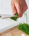 Hands using blue-handled scissors to cut fresh chives over a wooden cutting board.