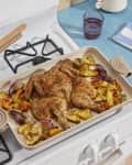 Roasted spatchcock chicken with pineapple, red onion, and peppers on a baking tray atop a stove, next to a plant and glass.